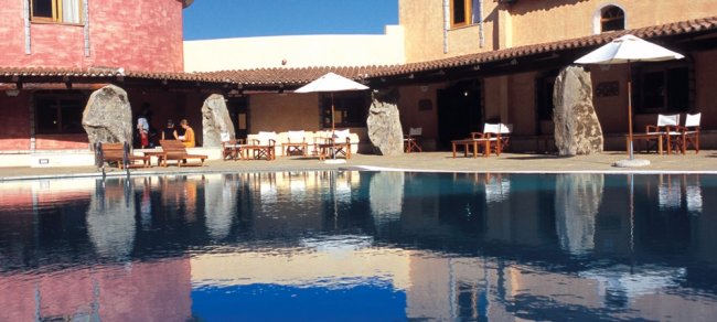 Piscina con sedie a sdraio e ombrelloni bianchi di fronte a un edificio con grandi rocce decorative.