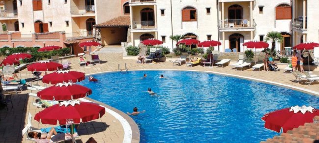 Una piscina con ombrelloni rossi e lettini, circondata da un edificio residenziale.