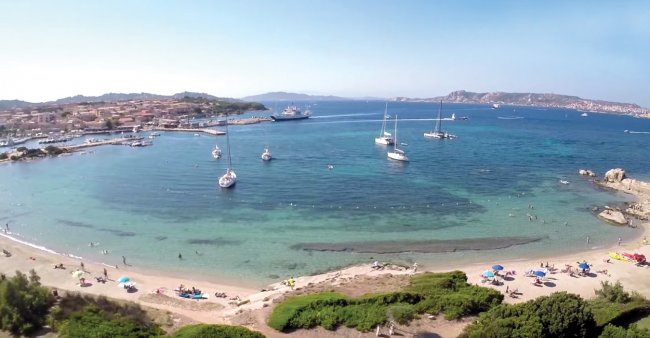 Spiaggia sabbiosa con barche a vela in mare e bagnanti sulla riva.